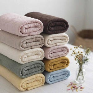 Op de afbeelding: Een stapel van acht gevouwen stoffen rollen in diverse gedempte kleuren, waaronder roze, donkerbruin, crème, lichtbeige, saliegroen, mosterdgeel en lichtblauw. De stof heeft een zichtbare gekreukte textuur. Een glazen vaas met droogbloemen staat ernaast.