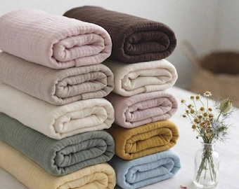May include: A stack of eight folded fabric rolls in various muted colours including pink, dark brown, cream, light beige, sage green, mustard yellow, and light blue. The fabric has a visible crinkled texture. A glass vase with dried flowers sits nearby.
