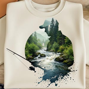 May include: Cream-colored sweatshirt featuring a silhouette of a person wearing a cap, overlaid with a scenic river and forest design. A fishing rod extends from the design, with a dark blue splash effect at the bottom. The design evokes a theme of fishing and nature.