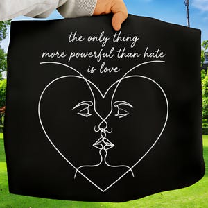 May include: Black tote bag featuring a white line drawing of two faces merging into a heart shape. The text "the only thing more powerful than hate is love" is above the heart design.