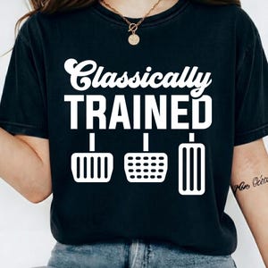 May include: Black t-shirt with the text "Classically TRAINED" in white, above three white pedal graphics. The shirt is a dark color and appears to be made of soft material.
