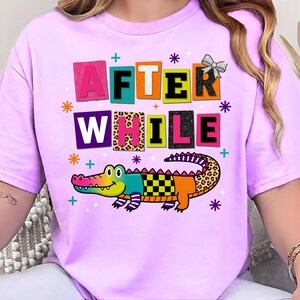 Puede incluir: Camiseta de color lila claro con la frase "AFTER WHILE" en letras de bloque coloridas y un cocodrilo de dibujos animados. El cocodrilo tiene un diseño de patchwork con varios patrones y colores, incluyendo estampado de leopardo y cuadros. La camiseta tiene cuello redondo.