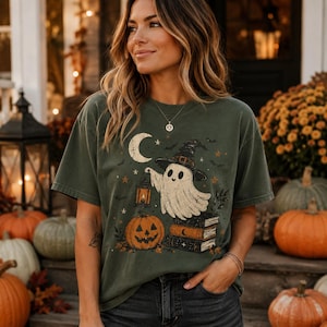 May include: A dark olive green short-sleeve t-shirt featuring a graphic print of a white ghost wearing a witch hat, holding a lantern, sitting on a stack of books next to a jack-o'-lantern, with a crescent moon, stars, and bats in the background.