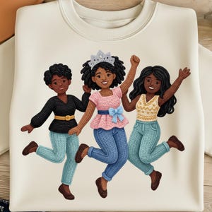 May include: Cream-coloured sweatshirt with a graphic of three smiling Black women in knitted outfits. The women are jumping and wearing different tops, including a black top with a gold belt, a pink top with a blue bow, and a yellow top.