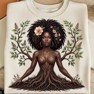 May include: Cream-colored sweatshirt featuring a digital illustration of a woman with tree-like features. Her body is depicted as a tree trunk and roots, with branches extending from her arms. She has dark hair adorned with flowers and wears gold hoop earrings.