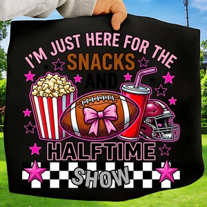 May include: Black graphic with a football, popcorn, drink, and helmet. Text says "I'M JUST HERE FOR THE SNACKS AND HALFTIME SHOW" in pink and white. Pink stars and a checkered pattern complete the design.