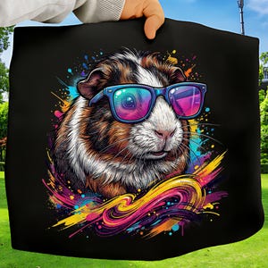 May include: Black tote bag featuring a colorful illustration of a guinea pig wearing sunglasses. The guinea pig has brown, white, and black fur. The design includes vibrant splashes of yellow, pink, blue, and purple.