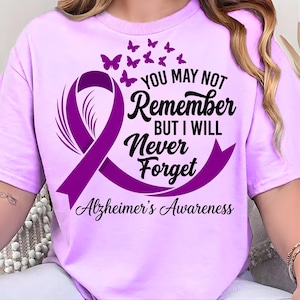 May include: A light purple t-shirt featuring a purple ribbon and butterflies, with the text "You May Not Remember But I Will Never Forget Alzheimer's Awareness". The shirt is made of a soft material.