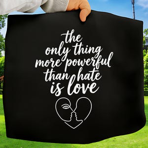 Peut inclure: Sac fourre-tout noir avec le texte blanc "la seule chose plus puissante que la haine est l'amour". Un dessin au trait blanc de deux visages formant un cœur est en dessous. Le sac est tenu par une personne.