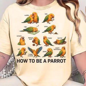 Puede incluir: Camiseta de color beige claro con un gráfico colorido de loros que demuestran varios comportamientos. El texto "HOW TO BE A PARROT" está impreso en negro en la parte inferior de la camiseta.