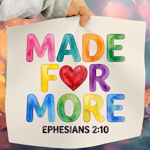Puede incluir: Un letrero de papel blanco sostenido por una mano muestra las palabras "MADE FOR MORE" en letras de acuarela coloridas. Un corazón rojo reemplaza la palabra "LOVE". El texto "EPHESIANS 2:10" está impreso en la parte inferior.