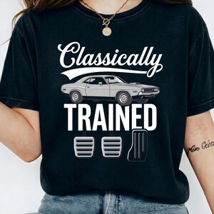 Może przedstawiać: Czarny t-shirt z białym napisem "Classically Trained", nad którym znajduje się szara ilustracja samochodu. Poniżej tekstu znajdują się ilustracje pedałów samochodowych. Koszulka jest wykonana z miękkiego materiału.