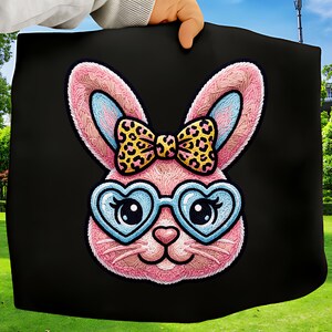 Könnte beinhalten: Ein schwarzes Stoffstück mit einem rosa Hasengesicht mit blau umrandeten herzförmigen Brillen und einer Schleife mit Leopardenmuster. Der Hase hat rosa Ohren mit blauen Innenohren. Das Bild wird von einer Hand gehalten.