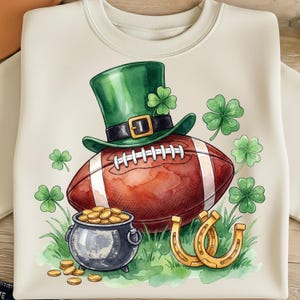 Może przedstawiać: Kremowa bluza z nadrukiem z okazji Dnia Świętego Patryka. Zielony kapelusz krasnoludka ze złotą klamrą spoczywa na piłce do futbolu amerykańskiego. Garnek złota, koniczyny i złote podkowy dopełniają świąteczną ilustrację.