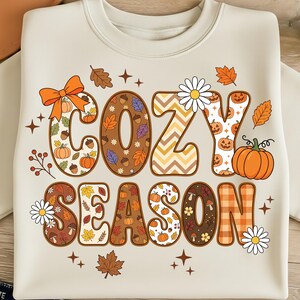 May include: Cream-coloured sweatshirt featuring the words "COZY SEASON" in a seasonal design. The letters are decorated with pumpkins, leaves, and floral accents. An orange bow and a pumpkin are also featured.