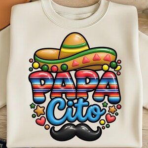 以下が含まれることがあります： クリーム色のスウェットシャツ。カラフルなグラフィックデザインで、「PAPA Cito」の文字がストライプで描かれ、ソンブレロ、黒い口ひげ、星やハートなどの装飾要素が特徴です。