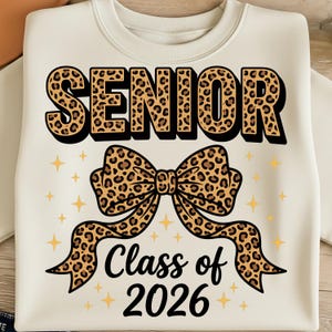 Può includere: Felpa color crema con la scritta "SENIOR" in grandi lettere con stampa leopardata. Sotto, un fiocco con stampa leopardata e il testo "Class of 2026". Accenti di stelle dorate sono sparsi intorno al design.