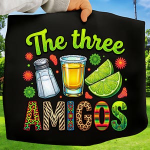 May include: Black graphic featuring a tequila shot, salt shaker, and lime wedges. The text "The three AMIGOS" is in green and colorful patterns. The image is held up by a person.