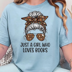 Könnte beinhalten: Hellblaues T-Shirt mit dem Text "JUST A GIRL WHO LOVES BOOKS". Die Grafik zeigt das Gesicht einer Frau mit einem Dutt und einer Sonnenbrille, beide mit einem Bücherregal-Muster gestaltet. Das Stirnband hat ebenfalls ein Bücherregal-Muster.