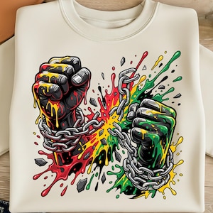 May include: Cream-colored sweatshirt featuring a graphic of two clenched fists breaking chains. The fists are black with yellow, red, and green accents, symbolizing unity and strength. The design includes splashes of color and shattered fragments.