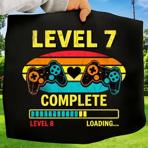 May include: Black graphic tee with the text "LEVEL 7 COMPLETE" in yellow and white. The design includes a retro sunset, two game controllers, a heart, and a loading bar. The words "LEVEL 8 LOADING..." are also present.
