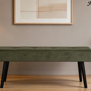 May include: An olive green upholstered bench with a corduroy texture. The bench has a rectangular shape with a button-tufted top and four black, angled legs. The bench is placed in front of a neutral-colored wall.