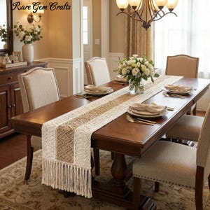 May include: A dining table set for a meal, featuring a cream and tan macrame table runner with fringe. The table is made of dark wood and surrounded by upholstered chairs. The room is decorated with flowers and a chandelier.