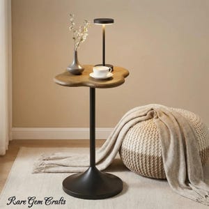 May include: A unique side table with a dark metal base and a wooden, cloud-shaped top. A small lamp, a cup and saucer, and a vase with flowers are on the table. A knitted pouf and a blanket are in the background. The table is approximately 28 inches tall.