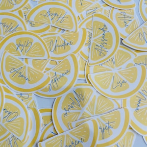 Peut inclure: Plusieurs autocollants en forme de quartiers de citron jaunes avec des noms manuscrits en bleu. Les autocollants ont une bordure blanche. Les noms incluent Christian, Muriel et Alessandra. Les autocollants sont éparpillés.