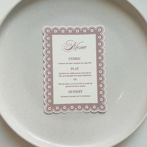 Peut inclure: Une carte de menu avec une bordure décorative, avec le mot "Menu" en écriture élégante. Le menu énumère une entrée, un plat principal et un dessert. La carte est placée sur une assiette grise clair mouchetée.