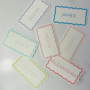 Peut inclure: Plusieurs cartons de noms avec des bords festonnés. Chaque carton présente un nom différent dans une couleur assortie, notamment "Elena", "James", "Julien", "Alicia", "Sophie", "Alexandra" et "Olivier". Les cartons sont sur une surface blanche.