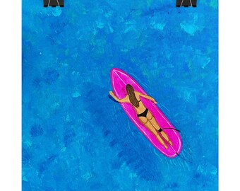 May include: A square art print featuring an overhead view of a person with long brown hair lying on a bright pink surfboard. The figure wears a black bikini. The background is a textured, vibrant blue surface representing deep ocean water.
