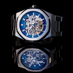 Könnte beinhalten: Eine silberfarbene Skelettuhr mit blauem Zifferblatt und sichtbaren Innenzahnrädern. Die Uhr hat ein geometrisches, achteckiges Gehäuse und ein passendes Metallarmband. Das Wort "THEMA" ist auf dem Zifferblatt aufgedruckt. Die Uhr spiegelt sich auf einer schwarzen Oberfläche.