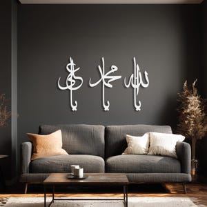 Könnte beinhalten: Weiße arabische Kalligraphie-Wandkunst. Das Kunstwerk zeigt drei separate Teile mit eleganter Schrift, die an einer dunkelgrauen Wand über einem grauen Sofa angebracht sind. Der Raum ist mit Kissen und einem Couchtisch aus Holz dekoriert.
