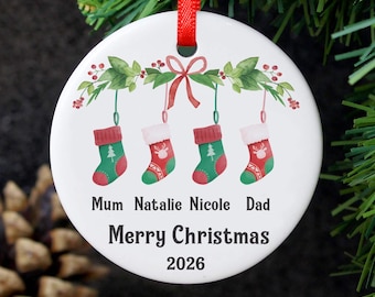 May include: A round white ceramic Christmas ornament hanging from a red ribbon. The design features four green and red stockings hanging from a leafy branch with a red bow. Text reads: Mum, Natalie, Nicole, Dad, Merry Christmas, and 2026.