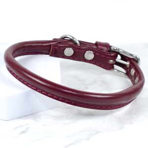 Puede incluir: Un collar de perro de cuero burdeos con un dise&ntilde;o redondeado. El collar presenta una hebilla plateada y dos remaches plateados. Los bordes est&aacute;n cosidos con un hilo rosa en contraste.
