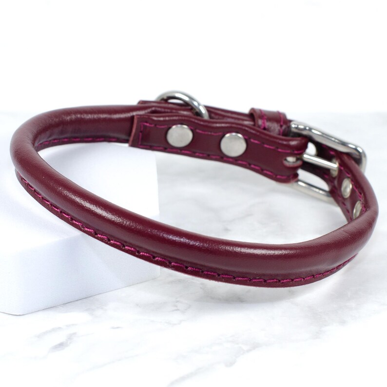 Puede incluir: Un collar de perro de cuero burdeos con un dise&ntilde;o redondeado. El collar presenta una hebilla plateada y dos remaches plateados. Los bordes est&aacute;n cosidos con un hilo rosa en contraste.