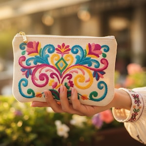 May include: A beige rectangular clutch with a gold zipper, featuring vibrant embroidery in pink, blue, yellow, and green. The embroidered design is a symmetrical floral pattern. The clutch is held in a person's hand.