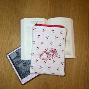 May include: A white quilted book sleeve with a red zipper and cherry pattern. The sleeve features two embroidered hearts with cherries and is next to an open book and an e-reader.