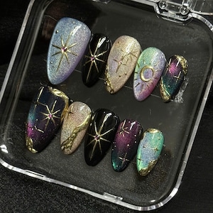 May include: A set of ten press-on nails featuring a celestial theme with cat-eye magnetic polish in shades of blue, purple, green, and black. The nails are decorated with gold metallic star, moon, and swirling line accents and small pink gems.