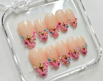 May include: A set of ten press-on nails featuring a nude base with pink French tips. Each nail is decorated with small, colourful translucent beads and gold-toned star-shaped charms, arranged in a clear plastic storage container.