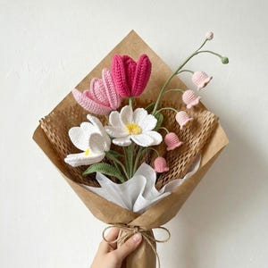May include: A bouquet of crocheted flowers featuring a deep pink tulip, a light pink tulip, two white daisies with yellow centres, and a stem of small, light pink bell-shaped flowers. The arrangement is wrapped in brown paper and white tissue.