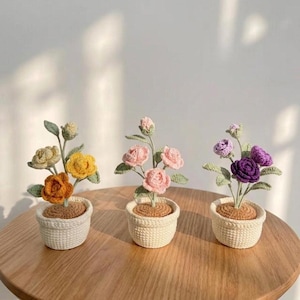 May include: Three crochet flower arrangements in white pots. The flowers are in shades of yellow, orange, pink, and purple, with green leaves. The pots sit on a wooden table, creating a charming, handmade aesthetic.