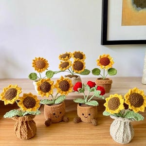 May include: Handmade crochet sunflowers and red flowers in small pots. The sunflowers have yellow petals and brown centres, with green leaves. The pots are various colours and styles, some with smiling faces.