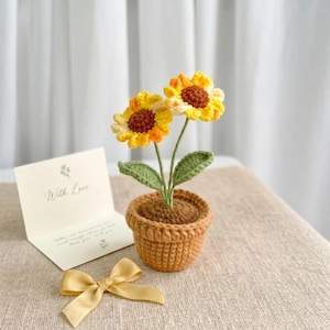 Op de afbeelding: Een gehaakte potplant met twee zonnebloemen met gele bloemblaadjes en bruine harten, groene bladeren en een bruine pot. Naast de pot ligt een klein crèmekleurig kaartje met de tekst "With Love" en een gele satijnen strik.