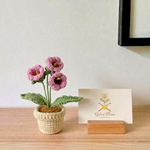 Peut inclure: Une plante en crochet en pot avec trois fleurs roses en forme de cloche aux centres jaunes et des feuilles vertes, dans un pot texturé couleur crème. À côté se trouve une carte rectangulaire blanche avec le texte Gifted Bloom CROCHET.