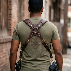 May include: A dark brown leather camera harness featuring a cross-back design with brass rivets, adjustable buckles, and metal swivel clips at the bottom to secure two cameras. The harness is worn over an olive green t-shirt with a graphic print.