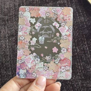 May include: A clear, rectangular resin piece with a skull image, surrounded by pink and white cherry blossoms, clouds, and small illustrations. A heart with the word "LOVE" is visible.