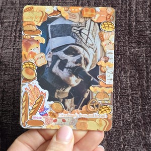 May include: A decorative card featuring a performer with skull-like makeup, surrounded by bread-themed stickers. The card includes the text "lucky Bread", "BREAD", and "Live, laugh, love, and bread."