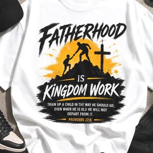 May include: A white short-sleeve t-shirt featuring a graphic of a person helping a child climb a mountain toward a cross. The text reads: FATHERHOOD IS KINGDOM WORK. TRAIN UP A CHILD IN THE WAY HE SHOULD GO; EVEN WHEN HE IS OLD HE WILL NOT DEPART FROM IT. PROVERBS 22:6.